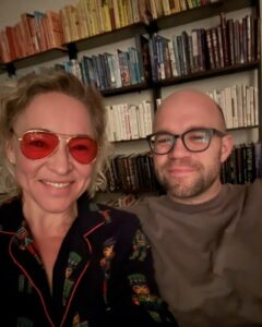 Kate in red-tinted glasses at home in the evening, in front of a wall of bookshelves.